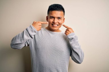 İzole edilmiş beyaz arka planda dikilen, gündelik süveter giyen, neşeli gülümseyen ve parmaklarıyla ve ağzıyla işaret eden genç Latin bir adam. Diş sağlığı konsepti.