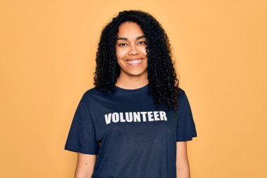 Genç Afro-Amerikalı kıvırcık kadın gönüllü olarak gönüllü olarak gönüllü tişört giyip mutlu bir yüz ifadesiyle ayakta duruyor ve gülümsüyor.