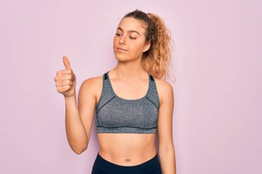 Genç, sarışın, mavi gözlü sporcu kadın spor kıyafeti giyiyor. Gururlu görünüyor, gülümsüyor. Kenarda baş parmak hareketi yapıyor.