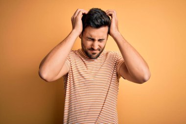 Sakallı genç yakışıklı adam sarı arka plan üzerine günlük çizgili tişört giyiyor. Ağrı ve migren yüzünden çaresiz ve stresli. Ellerini başının üstüne koy..