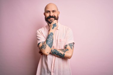 Sakallı ve dövmeli yakışıklı, pembe arka planda rahat gömlekli kolları çaprazlanmış ve elleri çenesinde kaldırılmış bir şekilde kameraya gülümseyen kel bir adam. Olumlu düşünüyorum..