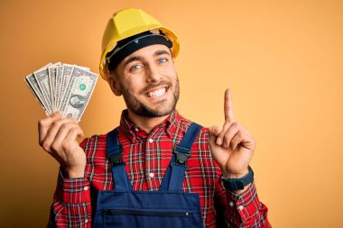 Güvenlik kaskı takan genç müteahhit, sarı arka plan üzerinde ödeme olarak dolar tutuyor. Bir fikir ya da soru işareti ile şaşırmış, mutlu yüzle bir numara.