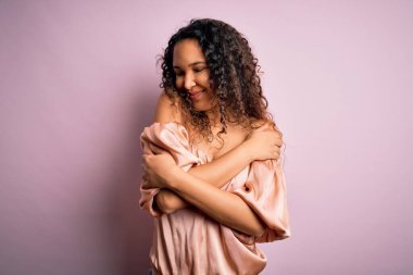 Kıvırcık saçlı, pembe tişörtlü genç ve güzel bir kadın pembe arka planda durup kendine sarılıyor, mutlu ve pozitif, kendinden emin gülümsüyor. Sevgi ve özgüven.