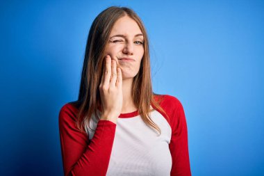 Genç, güzel, kızıl saçlı bir kadın diş ağrısı ya da diş ağrısı yüzünden izole edilmiş mavi arka plan üzerine günlük tişört giyip ağız ve acı dolu yüz ifadelerine dokunuyor. Dişçi