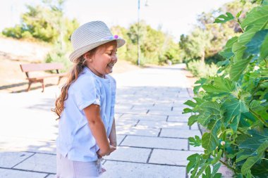 Yazlık şapkalı sevimli sarışın çocuk mutlu gülümsüyor. Yüzünde gülümsemeyle parkın etrafındaki yapraklara bakarak.