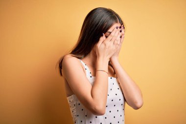 Sarı arka planda dikilen sıradan bir elbise giyen genç, güzel esmer kadın. Ağlarken yüzünü ellerle kapatan üzgün bir ifade. Depresyon kavramı.