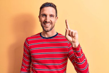 Sıradan çizgili tişört giyen genç yakışıklı adam izole edilmiş sarı arka planda duruyor. Bir fikir veya soruyla gülümsüyor ve mutlu bir yüzle işaret ediyor.