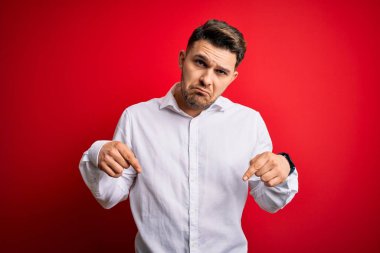 Mavi gözlü, zarif gömlekli genç bir iş adamı kırmızı izole edilmiş arka planda dikiliyor üzgün ve üzgün görünüyor, parmaklarıyla yönü gösteriyor, mutsuz ve depresif..
