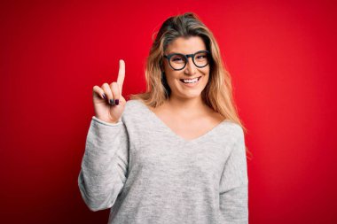 Genç, sarışın, süveter ve gözlük takan, izole edilmiş kırmızı arka planda bir numaralı parmağı gösteren, kendinden emin ve mutlu bir şekilde gülümseyen bir kadın..