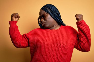 Sarı arka planda örgü ören Afro-Amerikan kadın kol kasları gururla gülümsüyor. Fitness konsepti.