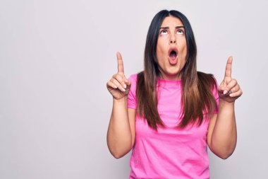 Genç, güzel, esmer, sıradan pembe tişörtler giyen bir kadın beyaz arka plan üzerinde şaşkın şaşkın duruyor ve parmaklarıyla işaret ediyor ve kollarını kaldırıyor..