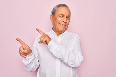 Üst düzey yakışıklı, gri saçlı, zarif gömlekli, pembe arka planda, gülümseyen ve kameraya bakan, iki eliyle ve parmaklarıyla yüzünü gösteren bir adam..