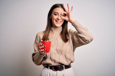 Genç, güzel, esmer bir kadın izole edilmiş beyaz arka planda kırmızı kahve içiyor. Gülümsüyor. Tamam.