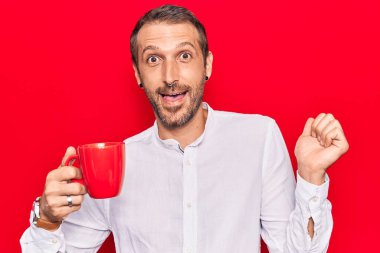 Genç yakışıklı adam elinde kahveyle gururla bağırıyor zaferi kutluyor ve başarıyı kollarını kaldırarak kutluyor. 