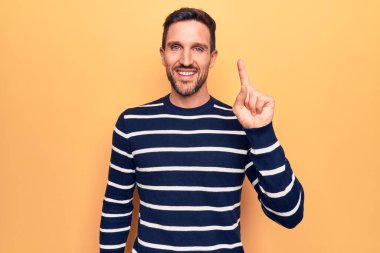 İzole edilmiş sarı arka planda dikilmiş sıradan çizgili süveter giyen genç yakışıklı bir adam bir fikir veya soruyla gülümsüyor ve mutlu bir yüzle işaret ediyor.