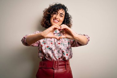 Güzel kıvırcık Arap kadın çiçek tişörtü giyip izole edilmiş beyaz arka planda duruyor. Sevgiyle gülümsüyor, elleriyle kalp sembolü yapıyor. Romantik konsept.