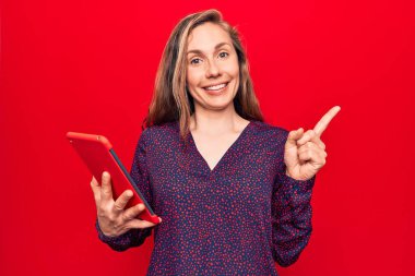 Sarışın, güzel bir kadın, izole edilmiş kırmızı arka plan üzerinde tablet kullanıyor. Gülümsüyor, mutlu bir şekilde işaret ediyor. 