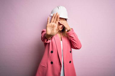 Genç ve güzel esmer mimar kadın pembe arka planda güvenlik kaskı takıyor gözleri ellerle örtüyor ve üzgün ve korku ifadeleriyle dur hareketi yapıyor. Utanç verici ve olumsuz bir kavram..