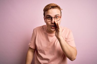 Genç, kızıl saçlı, günlük tişört giyen, izole edilmiş pembe arka planda duran ağzı açık, gizli dedikodular fısıldayan, kötü niyetli konuşmalar yapan genç bir adam.