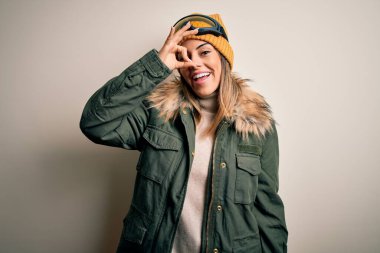 Genç esmer kayakçı kadın kar giysileri ve beyaz arka planda kayak gözlüğü takıyor. El hareketleriyle gülümsüyor, gözleriyle mutlu yüzüyle parmaklarından bakıyor..