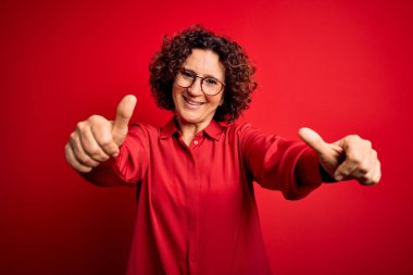 Orta yaşlı, kıvırcık saçlı, günlük gömlek ve gözlük takan güzel bir kadın. Kırmızı arka planda olumlu bir jest yapmayı onaylıyor. Elleriyle gülümsüyor ve başarı için seviniyor. Kazanan jest.
