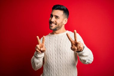 İzole edilmiş kırmızı arka planda dikilen beyaz süveter giyen yakışıklı genç adam kameraya bakıp zafer işareti yaparken gülümsüyor. İki numara..
