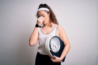 Beyaz arka planda tartı makinesi tutan kıvırcık saçlı genç bir kadın yorgun burnu ve gözleri ovuşturup yorgun ve baş ağrısı çekiyor. Stres ve hüsran kavramı.