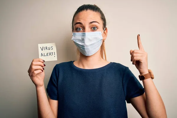 年轻美丽的女人 蓝眼睛 戴着医疗面具 在孤立的白色背景下 手持带有病毒警告信息的提醒纸站立 — 图库照片