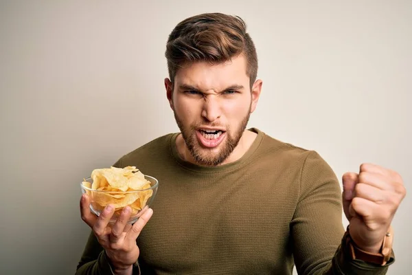 Young blond man with beard and blue eyes holding bowl with unhealthy potatoes chips annoyed and frustrated shouting with anger, crazy and yelling with raised hand, anger concept