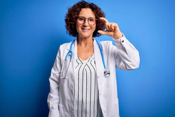 Middle age curly hair doctor woman wearing coat and stethoscope over blue background smiling and confident gesturing with hand doing small size sign with fingers looking and the camera. Measure concept.
