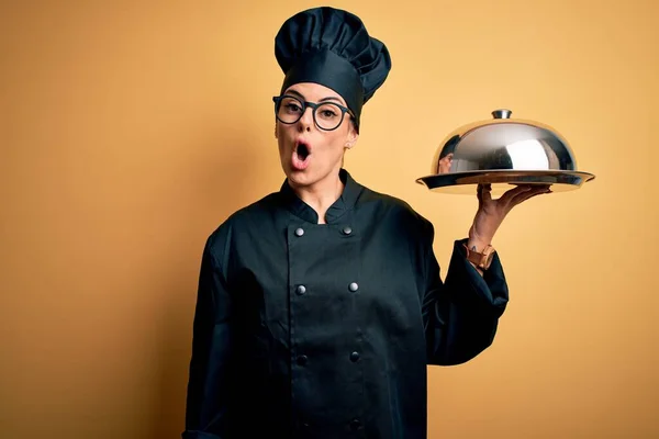 Young beautiful brunette chefwoman wearing cooker uniform and hat holding tray with dome scared in shock with a surprise face, afraid and excited with fear expression
