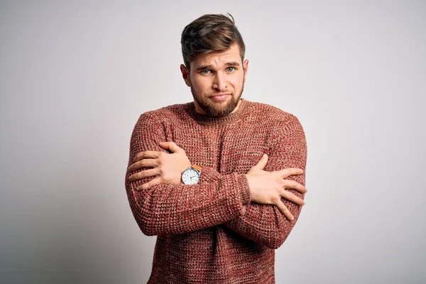 Young blond man with beard and blue eyes wearing casual sweater over white background shaking and freezing for winter cold with sad and shock expression on face