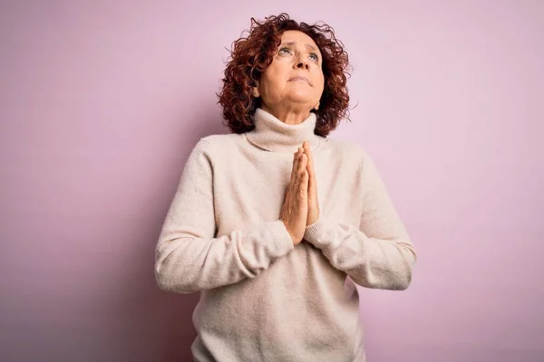 Middle age beautiful curly hair woman wearing casual turtleneck sweater over pink background begging and praying with hands together with hope expression on face very emotional and worried. Begging.