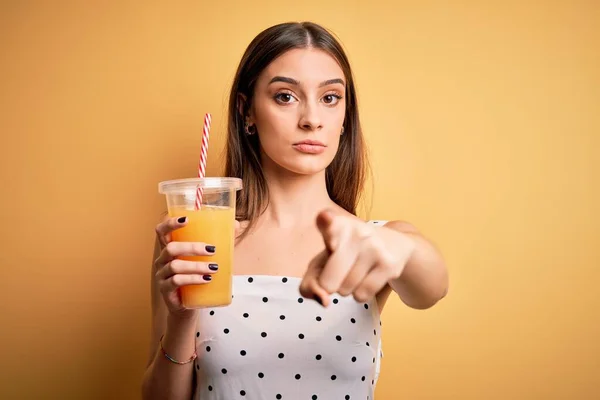 年轻美丽的黑发女子在黄色的背景下喝着健康的橙汁 手指指向摄像机和你 正面做出积极自信的手势 — 图库照片