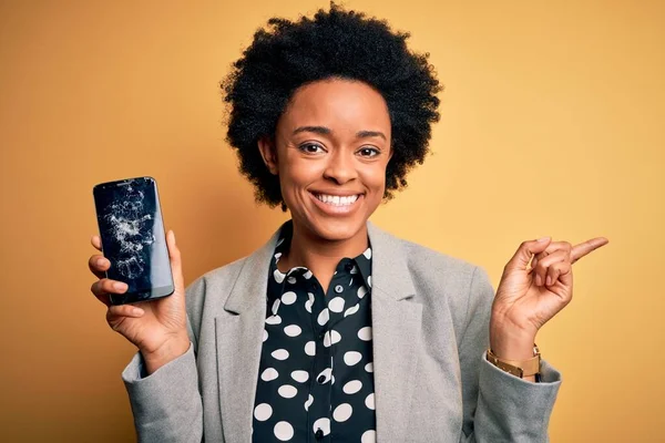 Young African American afro woman with curly hair holding cracked broken smartphone very happy pointing with hand and finger to the side