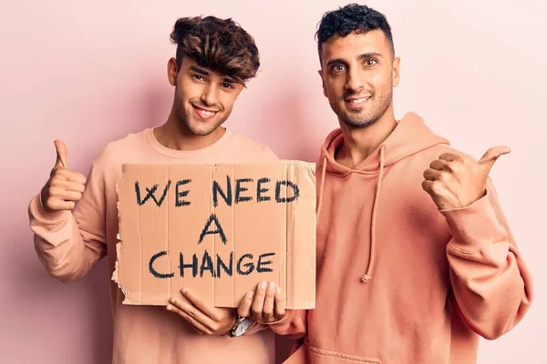 Young gay couple holding we need a change banner smiling happy and positive, thumb up doing excellent and approval sign