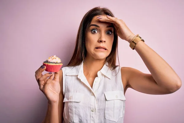 若い美しいブルネットの女性が孤立したピンクの背景の上にチョコレートカップケーキを食べて頭の上に手で強調し 恥と驚きの顔 怒りと不満でショックを受けた 恐怖と過ちへの動揺 — ストック写真