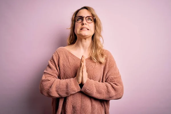 Young beautiful blonde woman wearing casual sweater and glasses over pink background begging and praying with hands together with hope expression on face very emotional and worried. Begging.