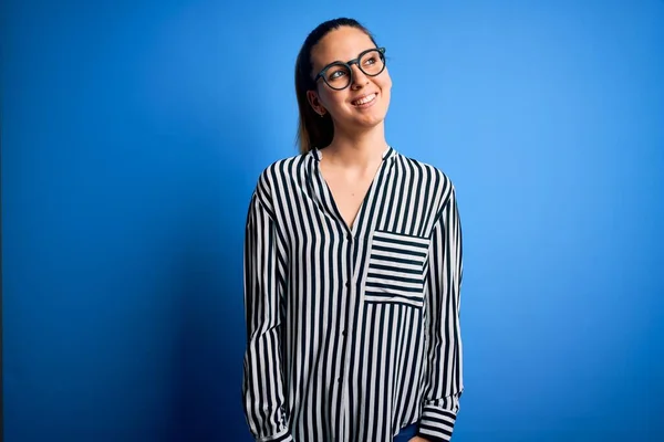 Beautiful blonde woman with blue eyes wearing striped shirt and glasses over blue background looking away to side with smile on face, natural expression. Laughing confident.