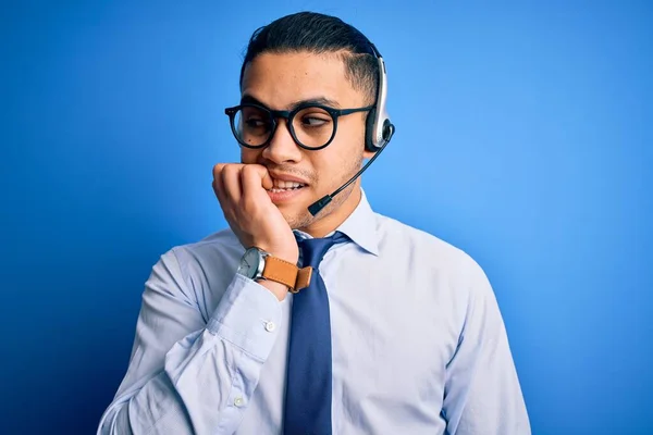 Young brazilian call center agent man wearing glasses and tie working using headset looking stressed and nervous with hands on mouth biting nails. Anxiety problem.