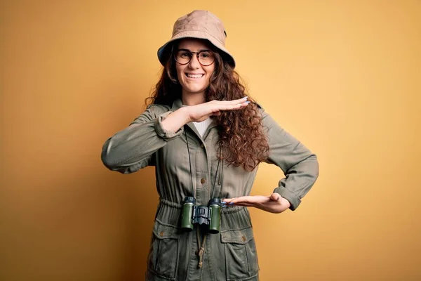 Young beautiful tourist woman on vacation wearing explorer hat and binoculars gesturing with hands showing big and large size sign, measure symbol. Smiling looking at the camera. Measuring concept.