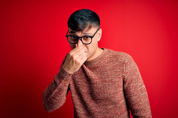 若いハンサムなヒスパニック系の男性は 鼻の上に指で息を保持し 臭いと嫌な 耐えられない臭い何かを嗅ぐ赤い背景の上にオタク眼鏡を着用 匂いが悪い — ストック写真