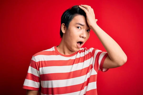 若いハンサムな中国人男性のカジュアルなストライプのTシャツを頭の上に手でミスのために エラーを覚えて驚いて赤い背景の上に立っている 忘れられた悪い記憶の概念 — ストック写真