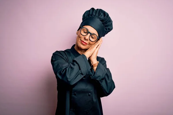 Middle age brunette chef woman wearing cooker uniform and hat over isolated pink background sleeping tired dreaming and posing with hands together while smiling with closed eyes.