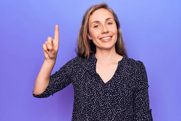 Genç, güzel, sarışın bir kadın, mor bir arka planda duruyor. Bir fikir veya soruyla gülümsüyor, mutlu bir yüzle işaret ediyor, bir numara. 