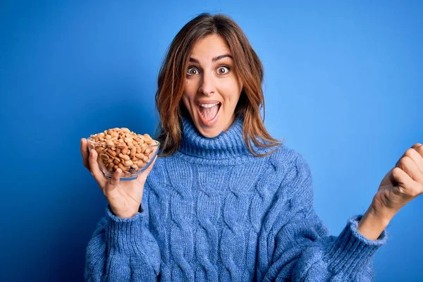 Young Beautiful Brunette Woman Holding Bowl Peanuts Blue Background Screaming — Stock Photo, Image