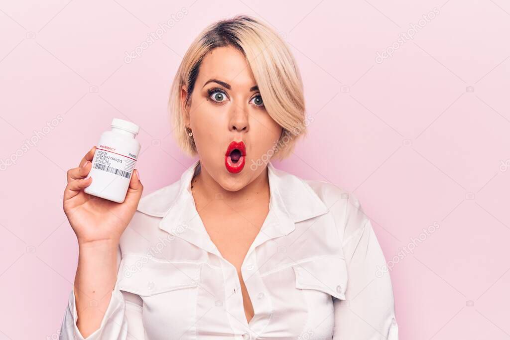 Young beautiful blonde plus size woman holding jar of pills over isolated pink background scared and amazed with open mouth for surprise, disbelief face