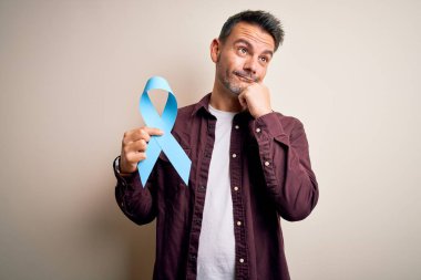 İzole edilmiş beyaz arka planda mavi kanser kurdelesi tutan yakışıklı genç adam soru soran ciddi bir yüz, kafası karışık bir fikir.