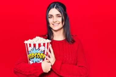 Genç ve güzel bir kız patlamış mısır yiyor. Pozitif ve mutlu görünüyor. Kendine güvenen bir gülümsemeyle ayakta duruyor ve dişlerini gösteriyor. 