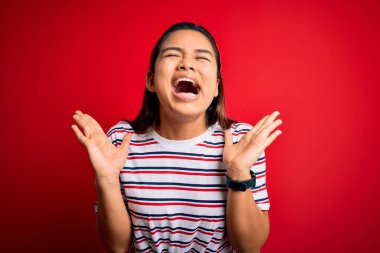 İzole edilmiş kırmızı arka plan üzerine sıradan çizgili tişört giyen genç, güzel Asyalı bir kız başarının çılgınlığını kollarını kaldırmış ve gözleri kapalı bir şekilde heyecanla haykırarak kutluyor. Kazanan konsept